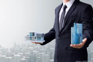 Mann im Anzug hält symbolisch ein modernes Einfamilienhaus und einen wolkenkratzerähnlichen Neubau in den Händen – Darstellung von Crowdinvesting im Immobilienbereich als Kapitalanlage zwischen Wohn- und Großprojekten.
